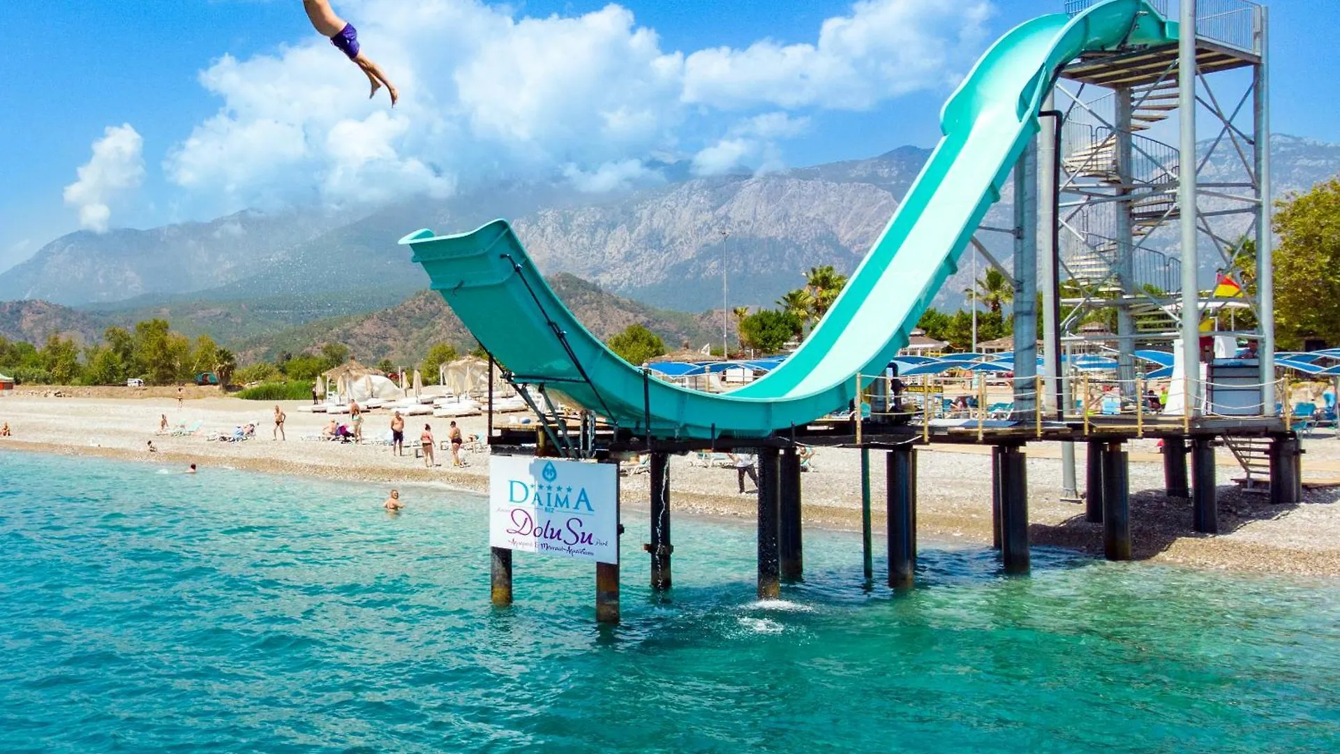 Daima Biz Hotel - Dolusu Aquapark Access Kemer Exterior photo This is a vibrant, sunny photo capturing a thrilling scene at a water park or resort.

The main focus is a **large, turquoise waterside slide** that curves gracefully into the bright blue water. The slide is supported by a metal platform structure bu